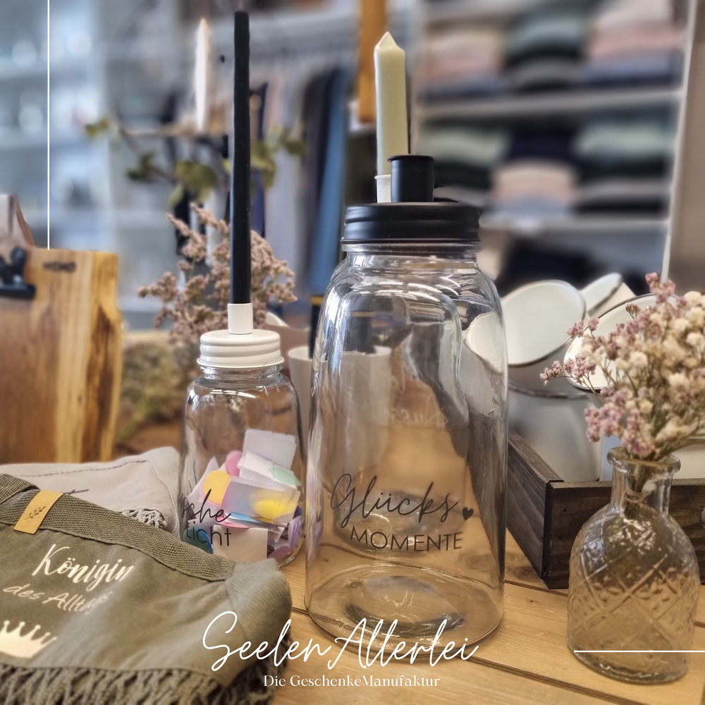 Kerzenglas mit Metalldeckel und der Aufschrift "Glücksmomente" in schwarz steht auf einem Tisch mit einem kleinen Kerzenglas mit weißem Deckel und einer schwarzen dünnen Stabkerze.