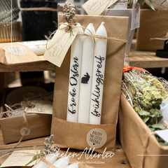 Stabkerzen mit anthrazitfarbener Schrift frühlingsrerif und Frohe Ostern stehen auf einem Holztisch