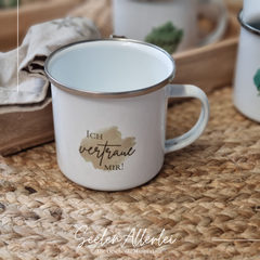 Emaille-Tasse mit silbernem Rand und der Aufschrift "Ich vertraue mit!" auf einem Holztablett
