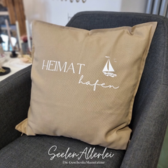 Beiges Sofakissen mit der Aufschrift "Heimathafen" und einem Segelboot liegt in einem Sessel
