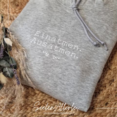 Grauer Hoodie mit Spruch Einatmen Ausatmen liegt auf Strohmatte mit Trockenblumen Dekoration von SeelenAllerlei