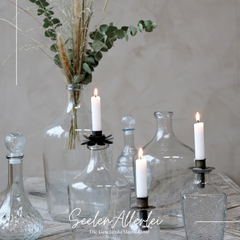 mehrere Glasvase und Glasflaschen stehtn auf einem holztisch. Eine Vase ist befüllt mit Trockenblumen, 3 verschiedene Kerzenhalteraufsätze für Flaschen stecken in drei verschiedenen Flaschen mit einer brennenden Kerze