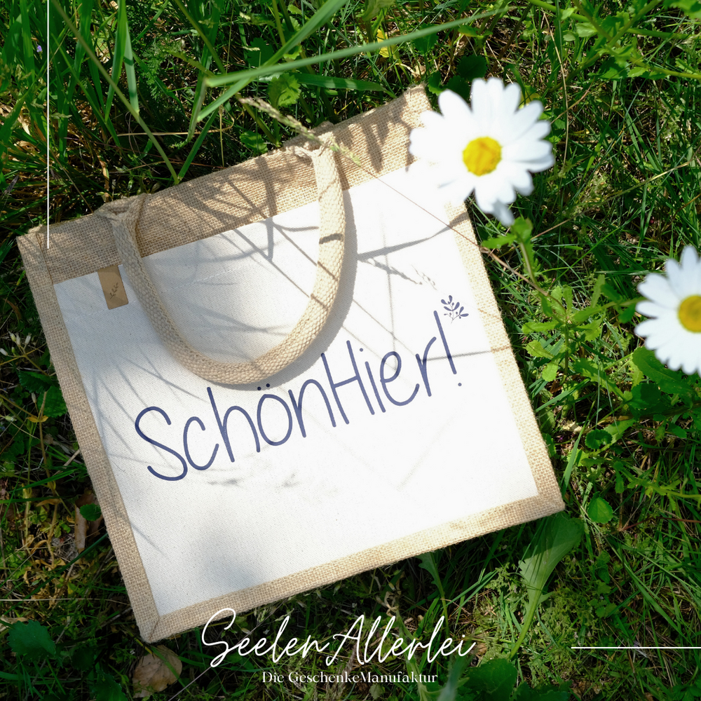 Eine Tasche mit der Aufschrift Schön Hier leigt im Gras mit Margeriten