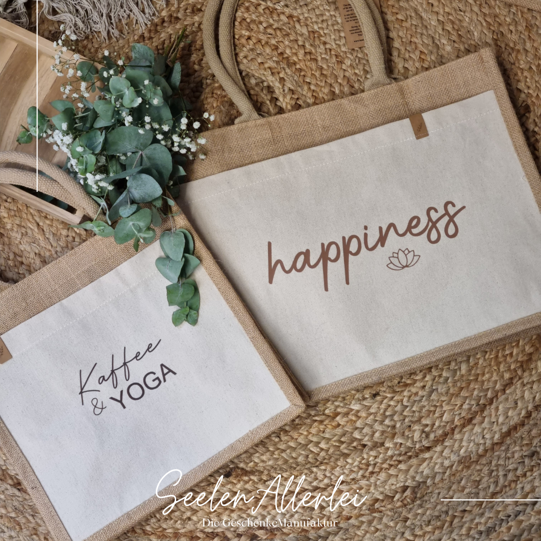 zwei Taschen in unterschiedlicher Größe und verschiedenen Aufdrucken liegen auf dem Boden. Auf der einen Tasche steht happiness auf der anderen Kaffee&Yoga