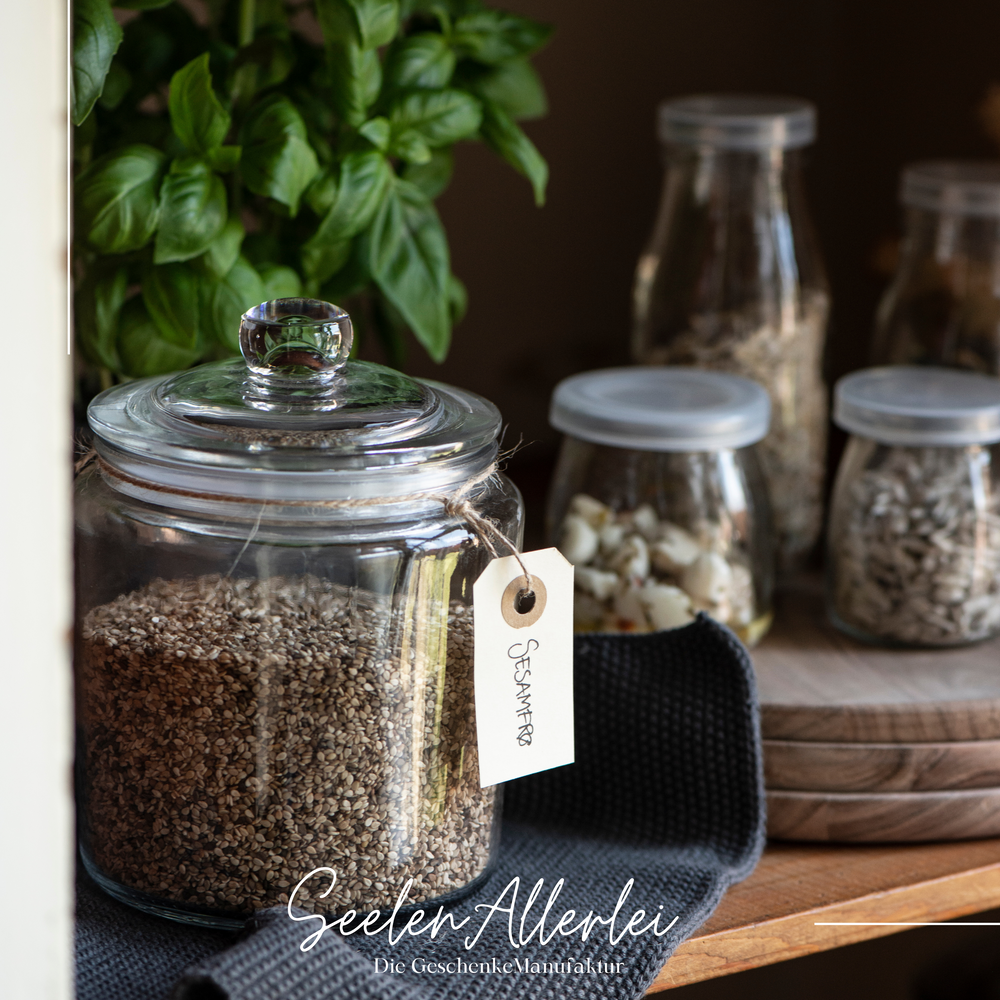 Ein Bonbonglas steht gefüllt mit Sesam in einem Regal mit weiteren Aufbewahrungsgläsern auf Holztabletts
