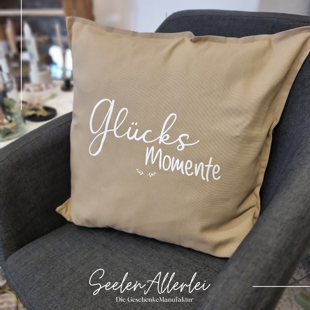 Kissen in beige mit der weißen Aufschrift Glücksmomente und zwei kleinen Zweigen  steht auf einem Sessel