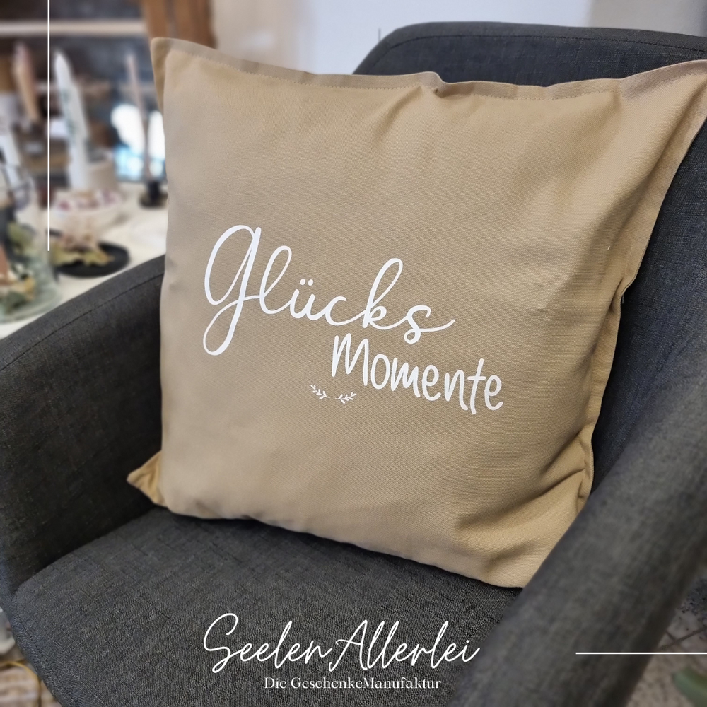 Kissen in beige mit der weißen Aufschrift Glücksmomente und zwei kleinen Zweigen  steht auf einem Sessel