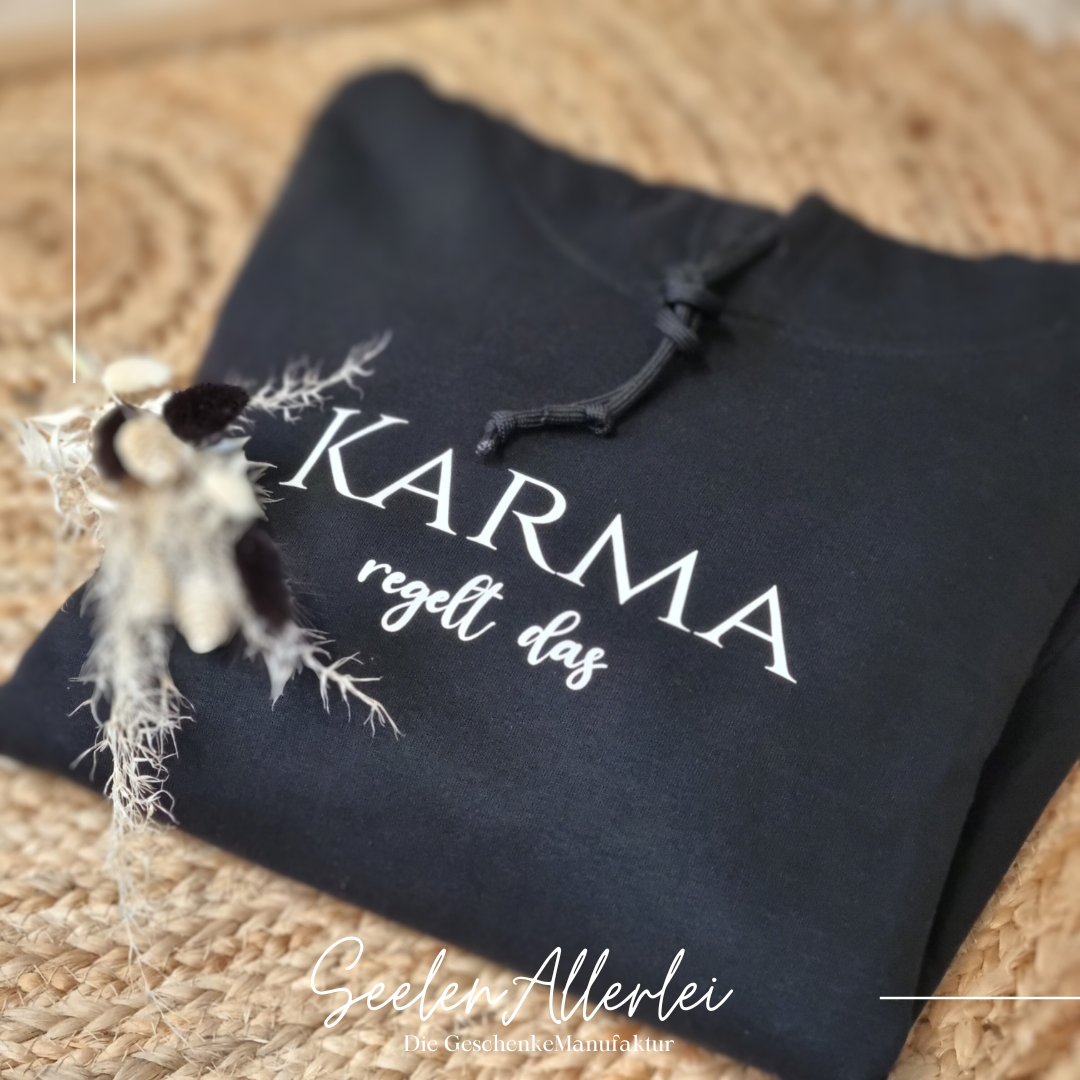 ein Pullover in schwarz mit der Aufschrift Karma regelt das in weißer Schrift liegt aufgefaltet auf dem Boden auf einem Teppich mit schöner Trockenblumendekoration