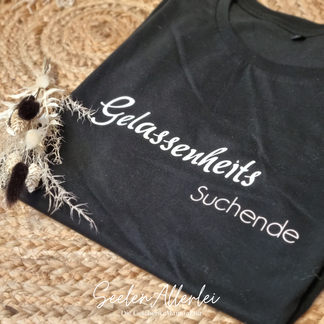 dunkelblaues T-shirt mit dem Spruch Gelassenheitssuchende liegt aufgefalten mit einer Trockenblume auf dem Teppeich