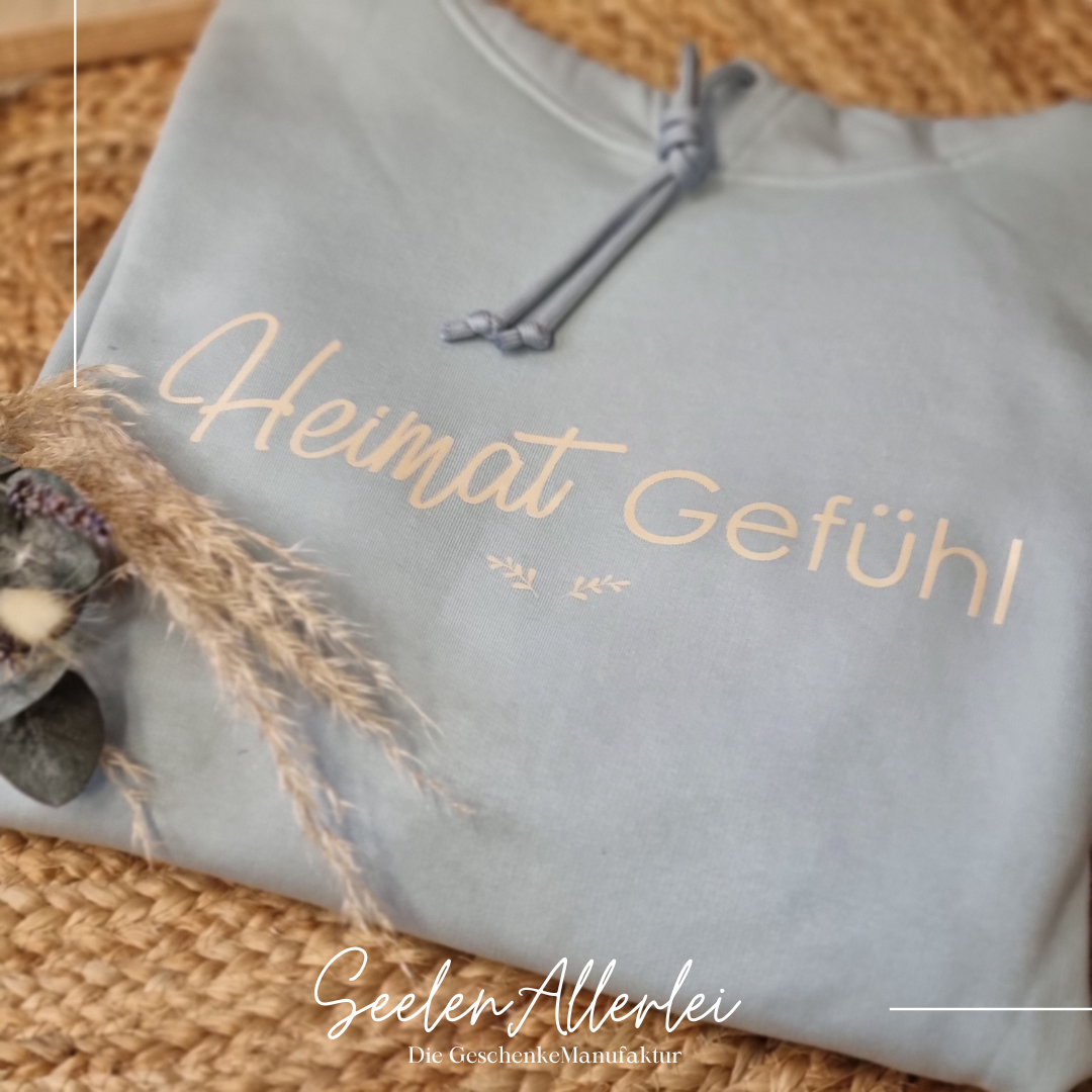 Heimatfefühl in beigefarbener Schrift steht auf einem hellblauen Hoodie, der Aufgefaltet auf dem Boden liegt