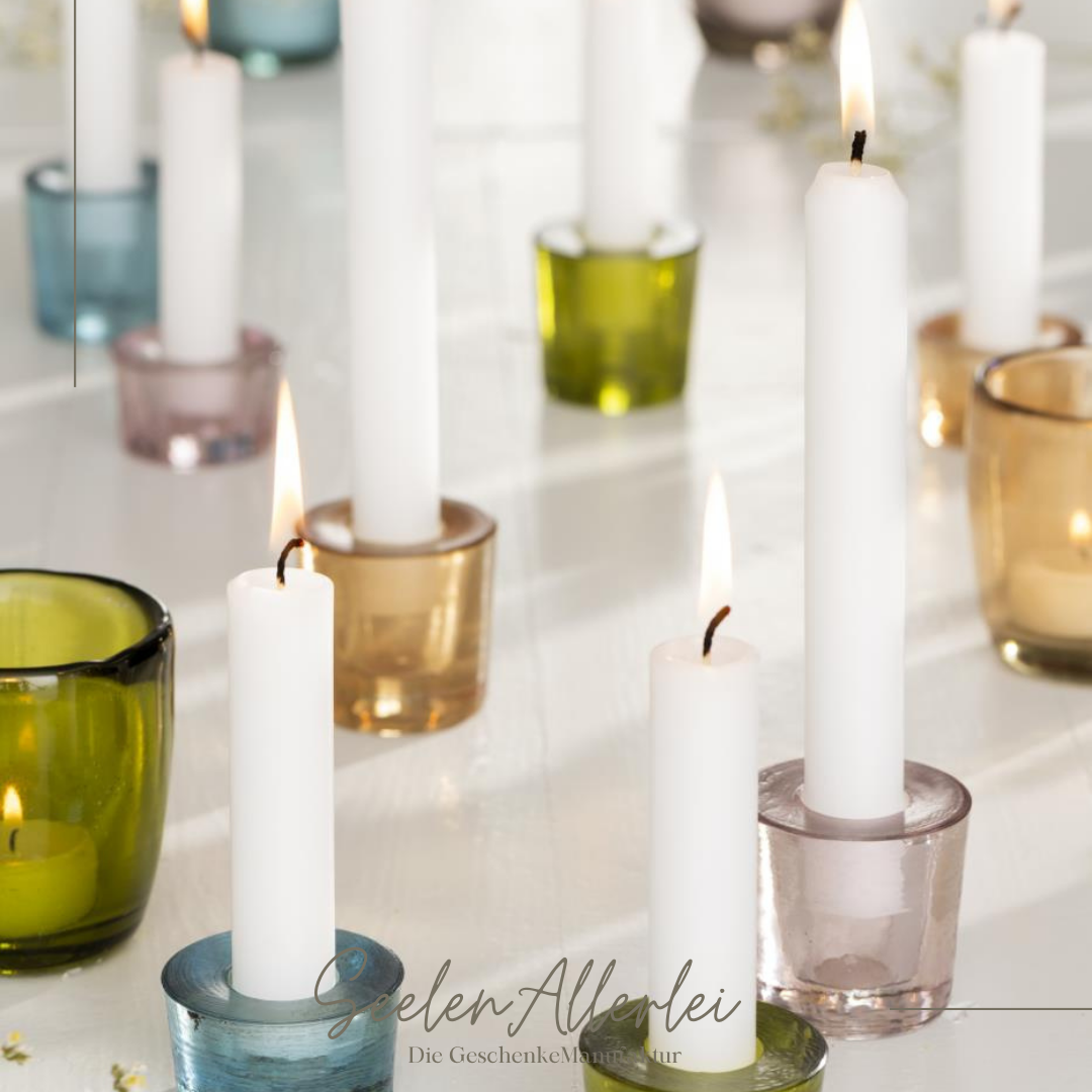 mehrere Kerzen stehen angezündet in verschiedenfarbigen Kerzenhaltern aus Glas auf einem Tisch