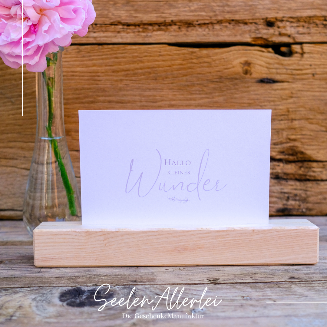 Hallo kleines Wunder steht auf einer weißen Postkarte, die in einem Kartenhalter aus Holz neben einer Blumenvase steht