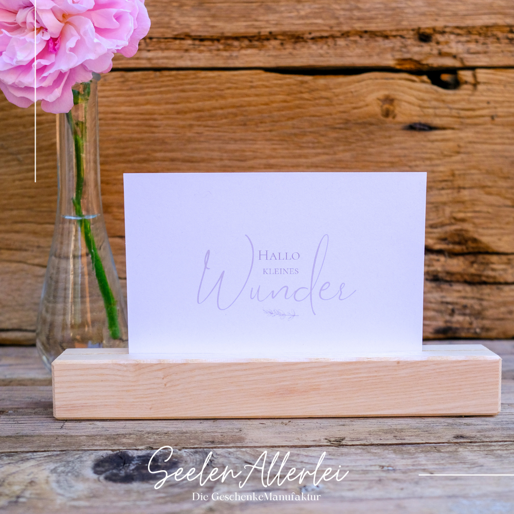 Hallo kleines Wunder steht auf einer weißen Postkarte, die in einem Kartenhalter aus Holz neben einer Blumenvase steht