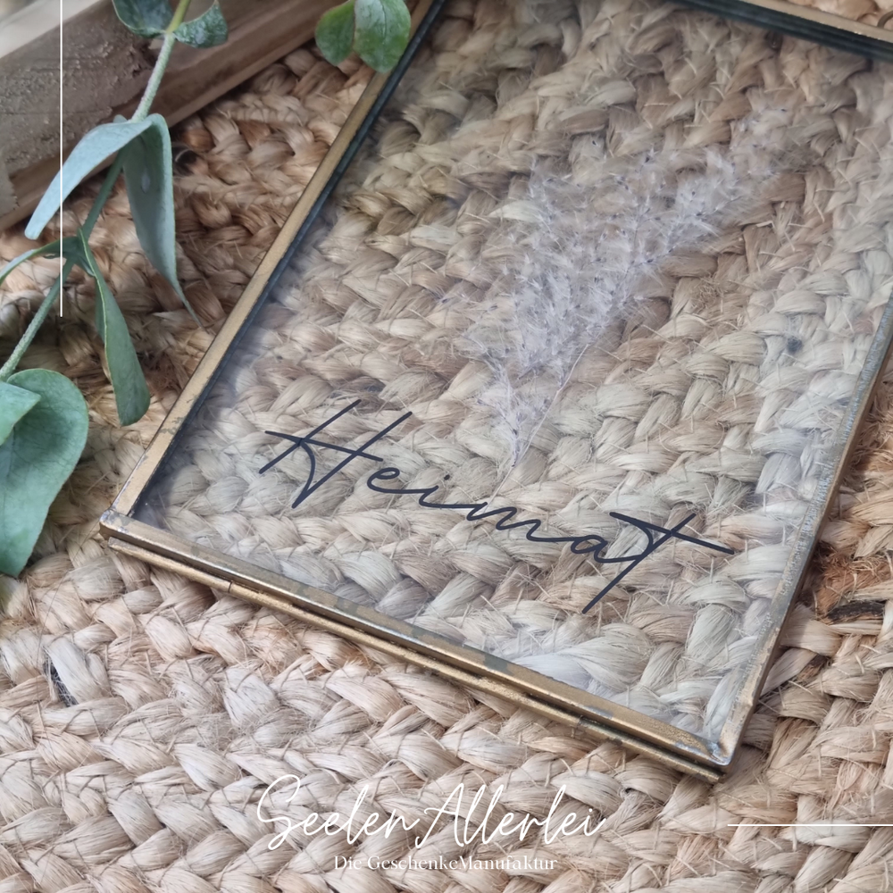 goldener kleiner Glasrahmen mit der Aufschrift Heimat liegt auf einem Korbteppich dekoriert