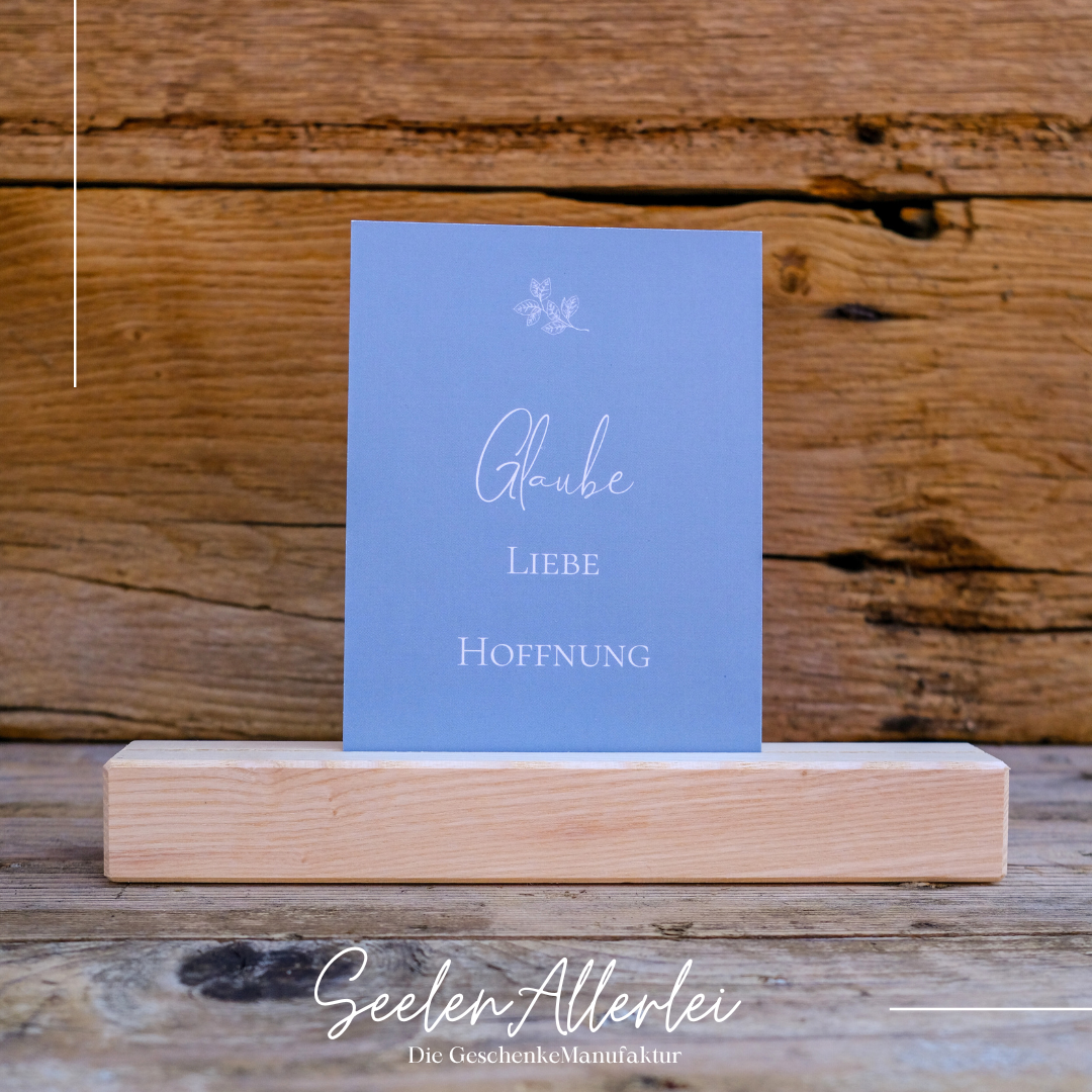 die Wörter Glaube  Liebe Hoffnung  stehen auf einer Glückwunschkarte in blau türkis, die in einem Kartenhalter aus Holz steht