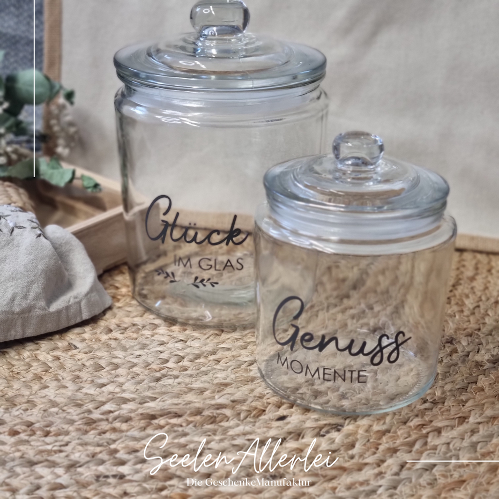 zwei verschieden Große Aufbewahrungsgläser stehen mit unterschiedlichen Aufschriften auf einem Teppich vor einer Markttasche und einem Holztablett