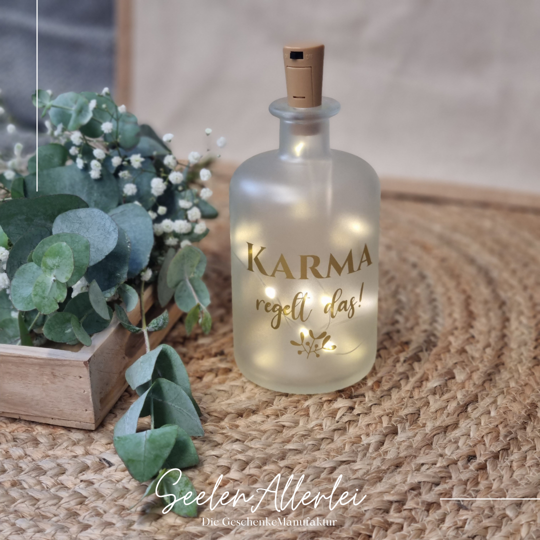 Karma regelt das steht in goldener Schrift auf einer Apothekerflasche mit Lichterkette steht auf einem Teppich mit Trockenblumendekoration