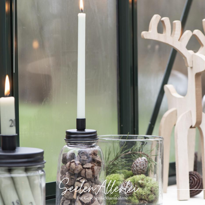 Kerzenhalterdose in klein für dünne Stabkerzen steht mit einer brennenden Kerze vor dem Fenster mit anderen Glasartikeln auf der Fensterbank