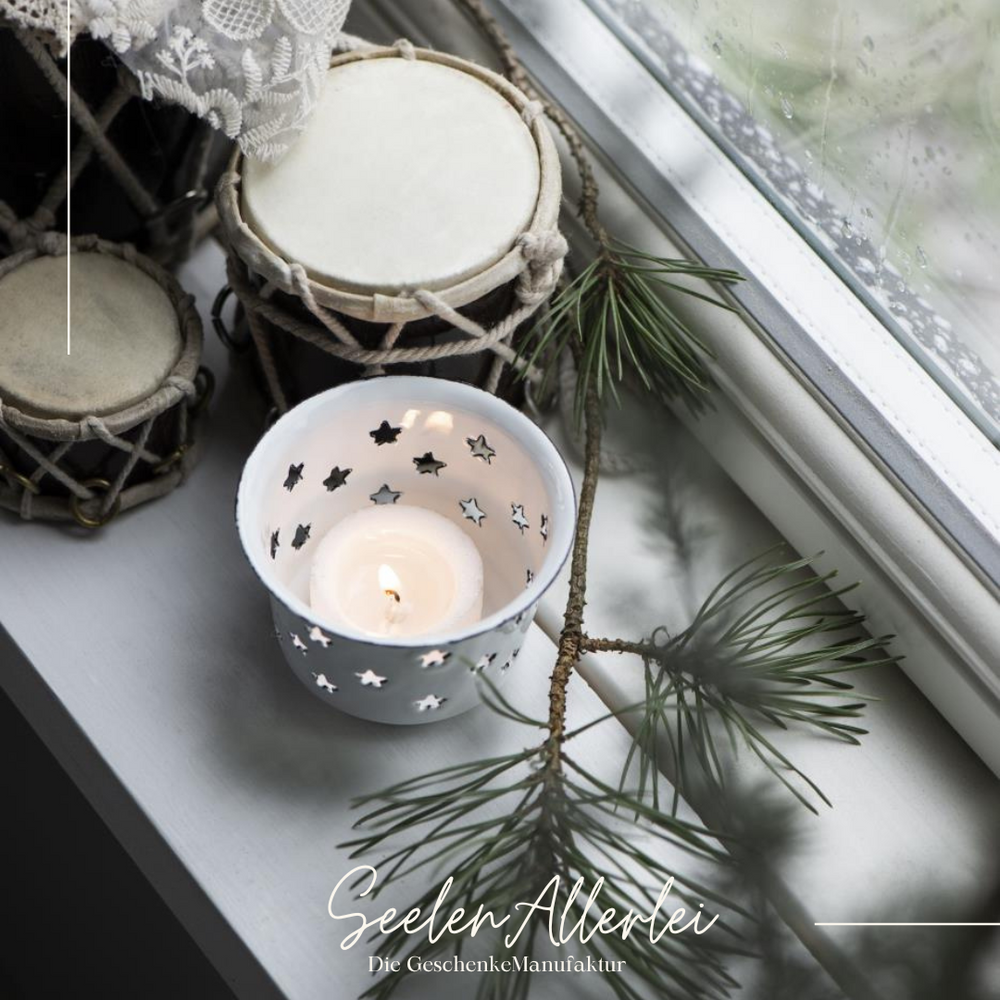 ein brennendes Teelicht steht im Kerzenhalter aus Emaille auf einer Fensterbank mit Zweigen und kleinen Trommeln