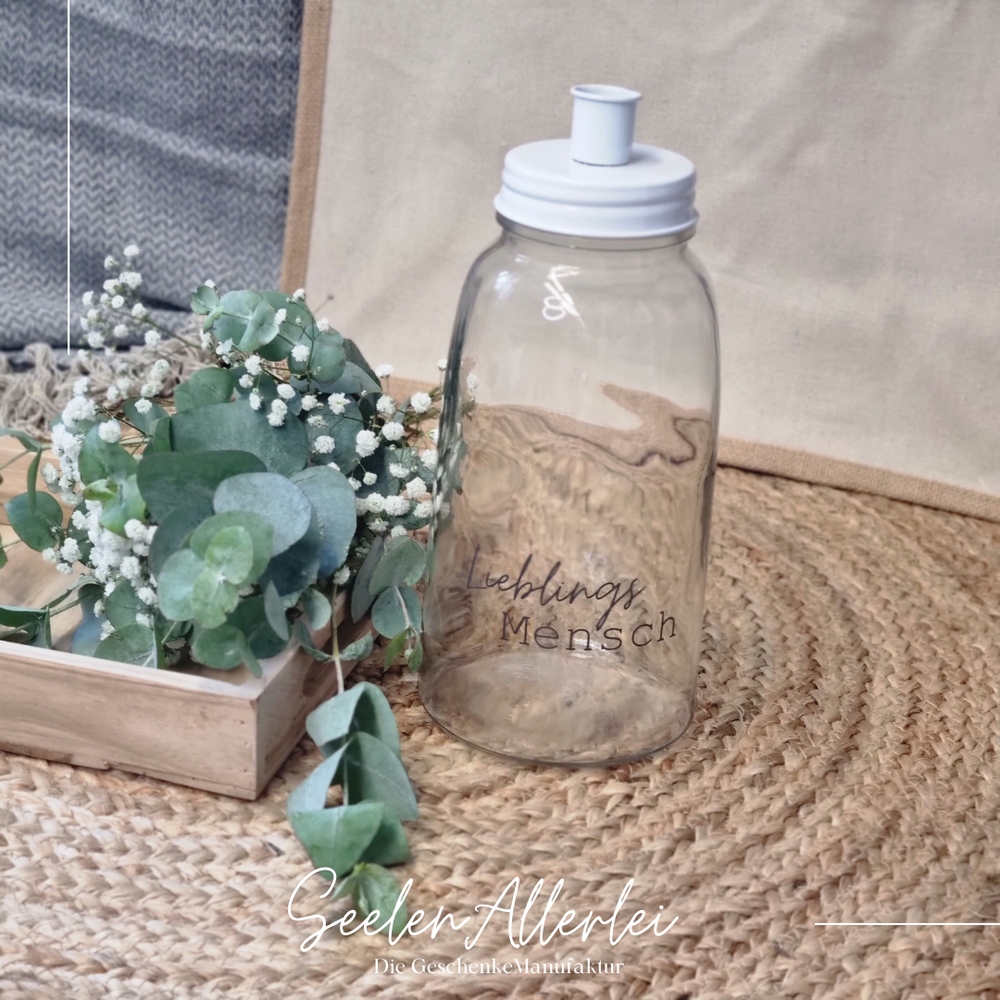 Kerzenlgas mit weißem Metalldeckel und der Aufschrift Lieblingsmensch steht auf einem Teppich mit Eukalyptusdekoration in einem Holztablett