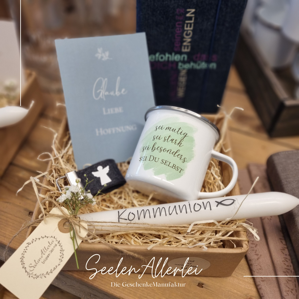 Geschenkschachtel zur Kommunion gefüllt mit einer Glückwunschkarte, einem Schlüsselanhänger mit Schutzengel, einer Kerze und einer Emaille-Tasse  vor einem Gebetbuch.