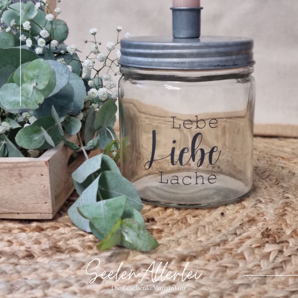 Lebe Liebe Lache steht in schwarzer Schrift auf einem Kerzenglas mit zinkfarbenen Metalldeckel