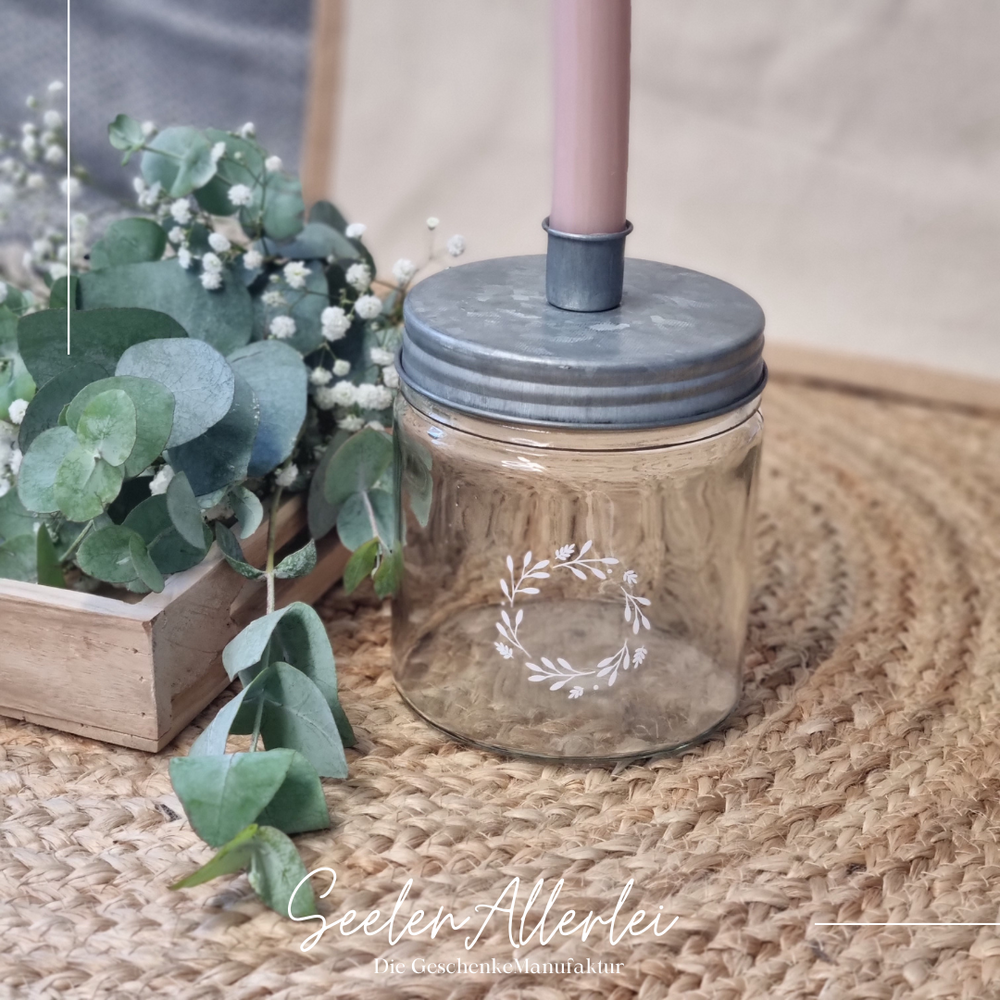 ein Lbumenkranz aus Zweigen ist auf einem Kerzenglas aufgedruckt, in der eine rosa Kerze steht