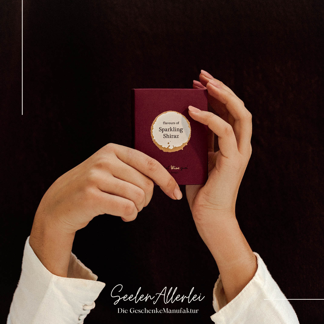 Produktfoto für die Variane Sparkling shiraz in der Wine gums verpackung von Vinoos im typishen Design. Nahaufnahme von Händen, die die Verpackung hochhalten vor dunkelrotem Hintergrund.
