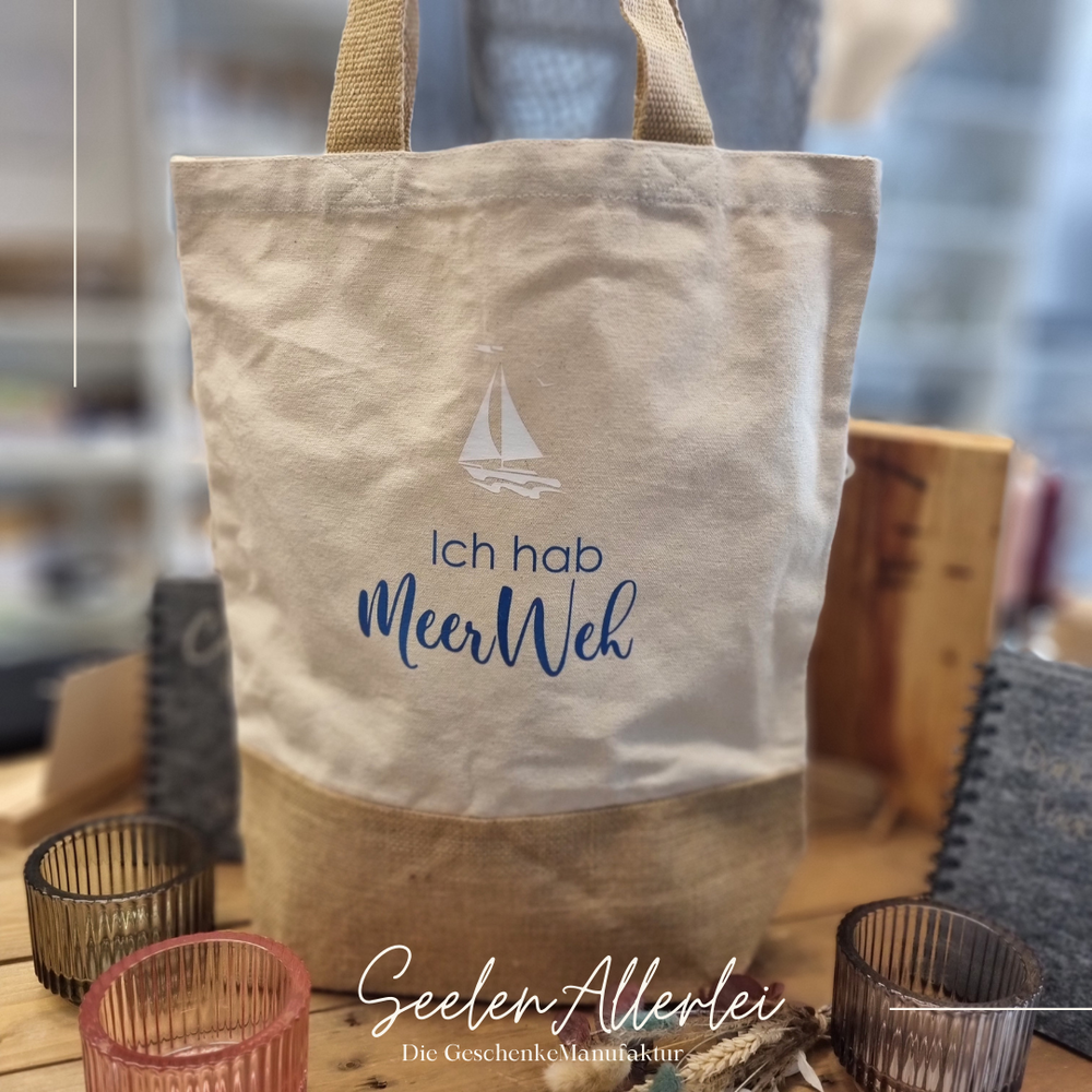 Ich habe Meerweh steht auf einem Shopper mit einem Segelboot in der Farbe weiß, die Tasche steht zusammen mit Teelichthaltern und Trockenblumen auf einem Holztisch