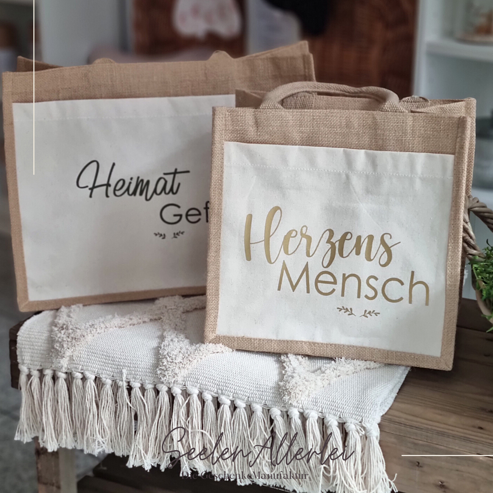 Beispielfoto für die Aufdrucke Herzensmensch und Heimatgefühl, beide Taschen stehen auf einer Bank mit einem Teppich drauf