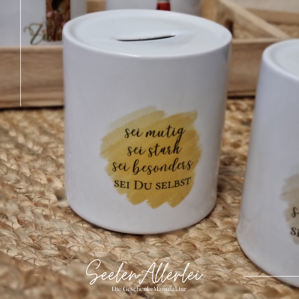 Spardose aus Porzellan mit dem spruch sei mutig sei stark sei besonders sei Du selbst steht neben einer anderen Spardose auf dem Teppich vor einem Tablett