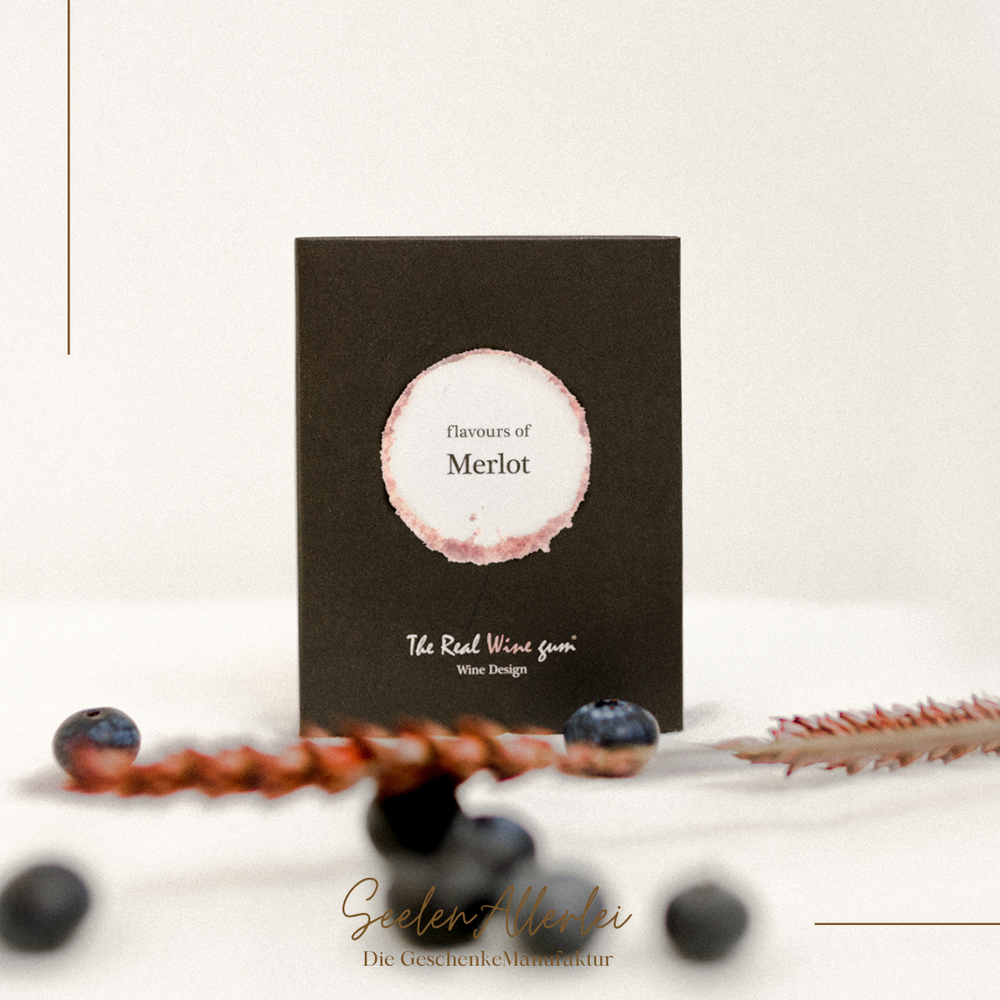 Merlot WineGums von Vinoos in schwarzer Verpackung als Nahaufnahme vor weißem HIntergrund  mit Blaubeeren als Dekoration. 