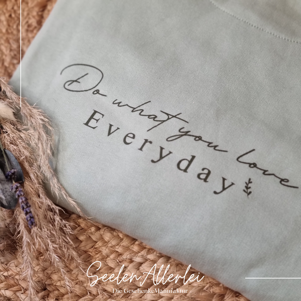 Weißer Hoodie mit schwarzem Aufdruck Do what you love Everyday liegt auf Strohmatte mit natürlicher Trockenblumen Dekoration