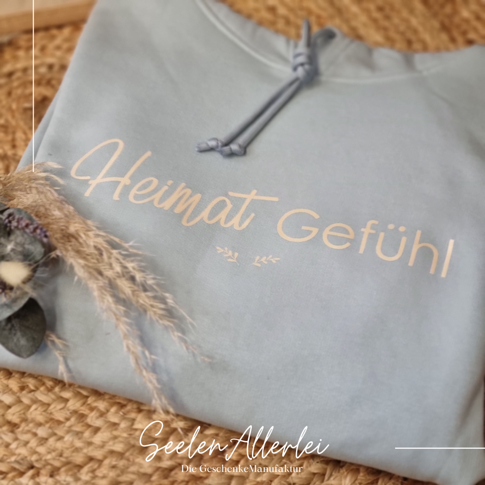 Hellgrauer Hoodie mit goldenem Aufdruck Heimat Gefühl liegt auf geflochtener Matte mit getrockneten Blumen als Dekoration