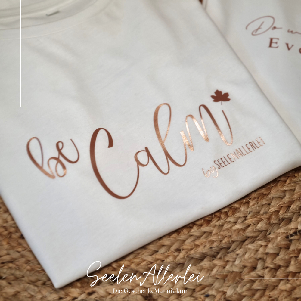 Weißes T-Shirt mit goldenem Aufdruck Calm liegt auf natürlichem Untergrund mit Trockenblumen und SeelenAllerlei Branding