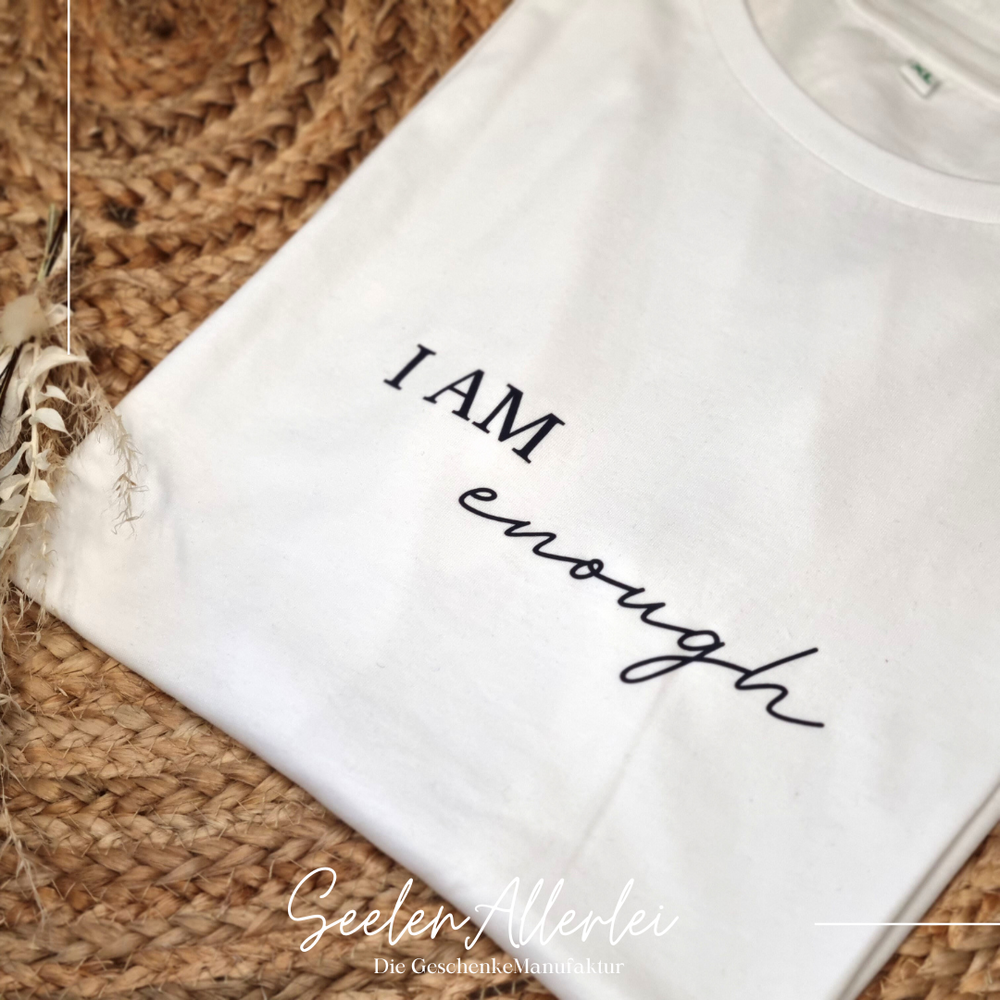 Weißes T-Shirt mit schwarzem Aufdruck I AM enough liegt auf geflochtener Matte mit natürlicher Trockenblumen Dekoration