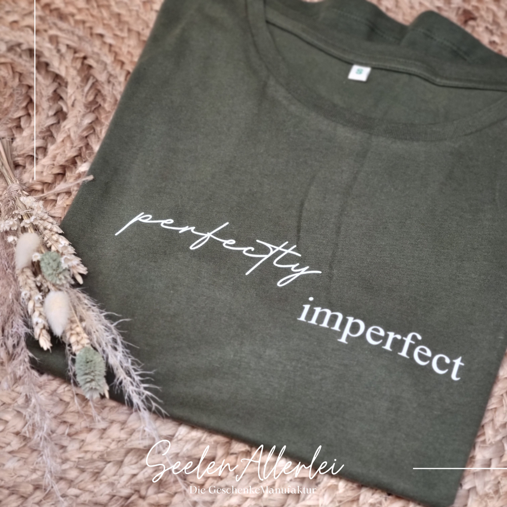 Dunkelgrünes T-Shirt mit weißem Aufdruck perfectly imperfect liegt auf Strohmatte mit getrockneten Blumen als Dekoration