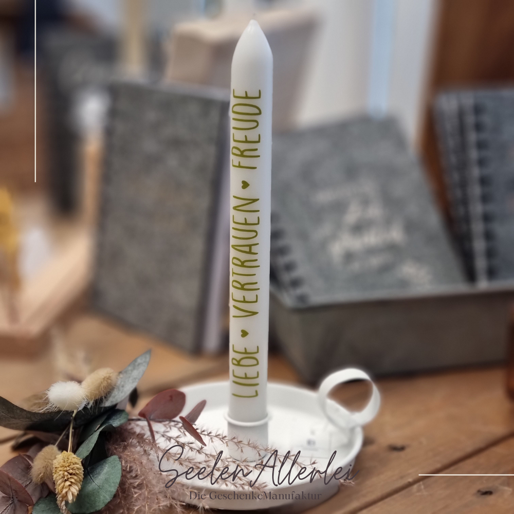 Stabkerze mit der Aufschrift Liebe - Vertrauen - Freude in einer Kammerleuchte in weiß, steht auf einem Holztisch vor einer Metallschale mit Trockenblumen